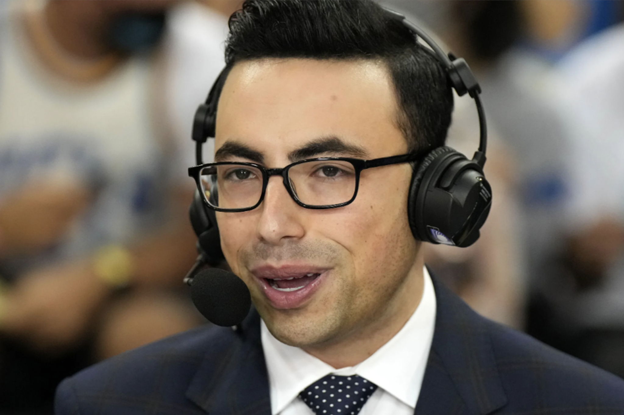 A sportscaster calls a game while wearing a suit, glasses and headset